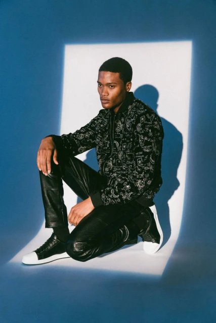 A young Black male model in a black patterned hoodie and shiny black pants sits on a blue background with a white spotlight.