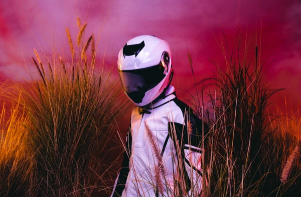 Person in white motorcycle jacket and helmet stands amid tall golden reeds against a vibrant purple and orange gradient background.