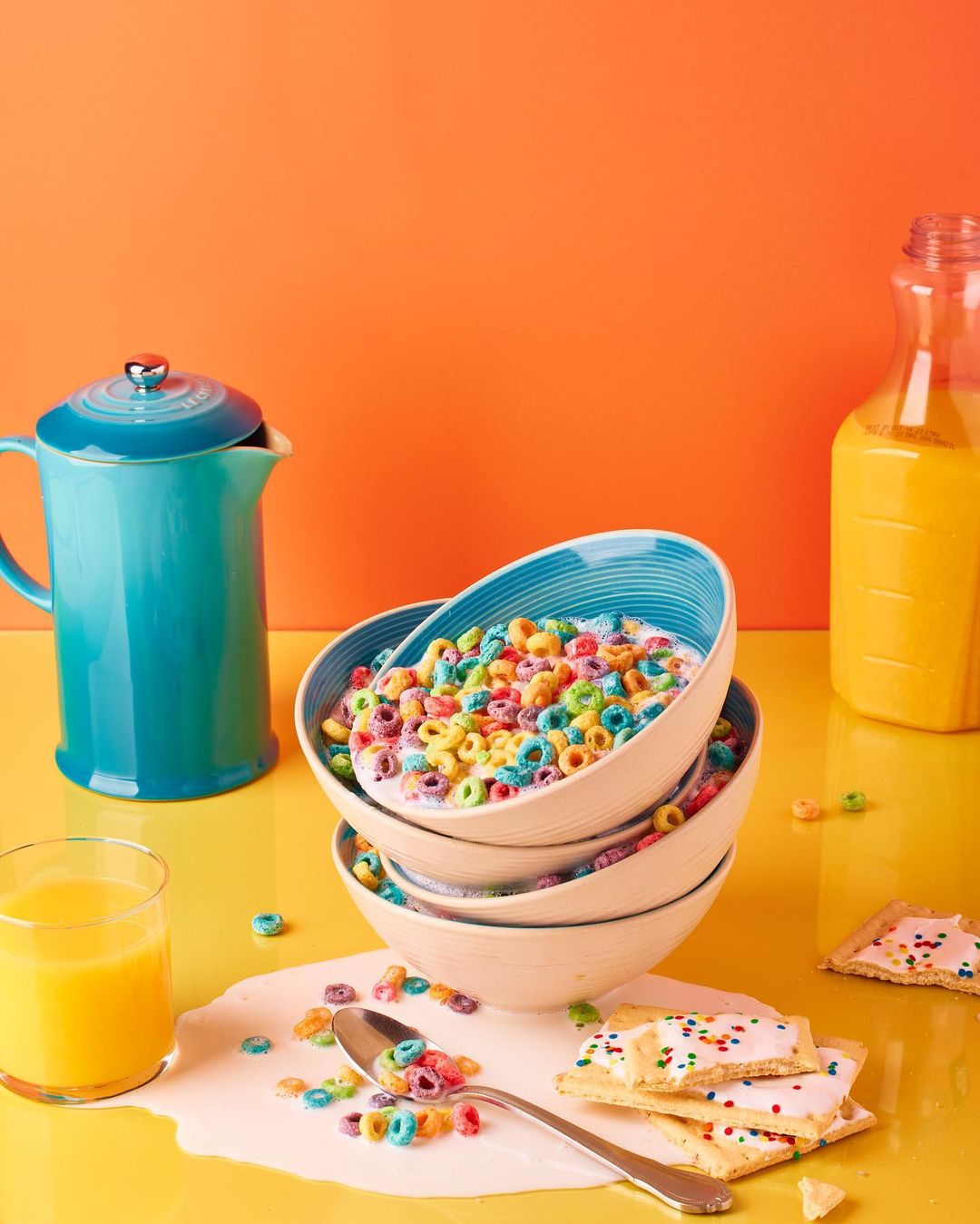 A vibrant still life featuring a stack of colorful cereal bowls, a blue pitcher, and a glass of orange juice against a bright orange background.