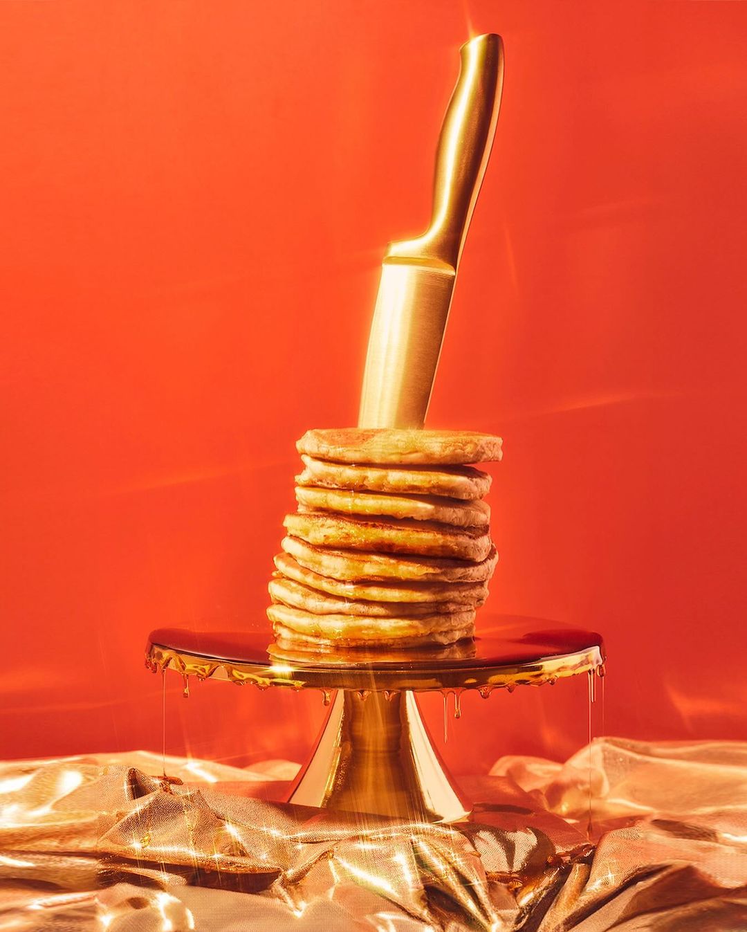 Stack of pancakes on a gold cake stand with syrup, stabbed by a gold knife, against a red background with shiny gold fabric.