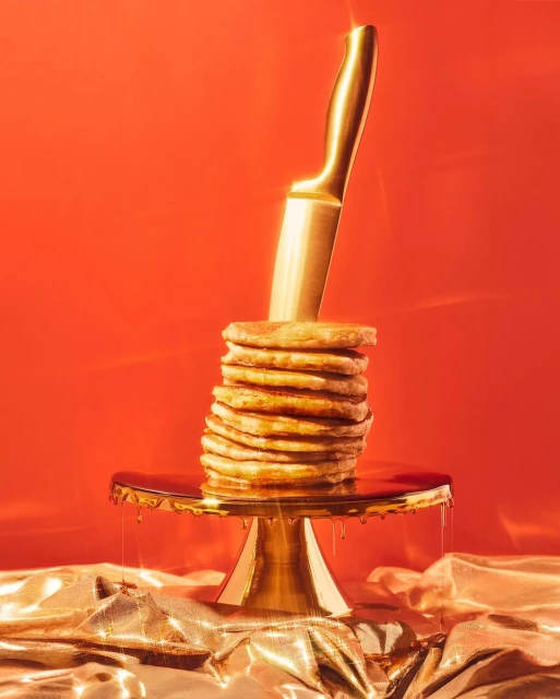 Stack of pancakes on a gold cake stand with syrup, stabbed by a gold knife, against a red background with shiny gold fabric.