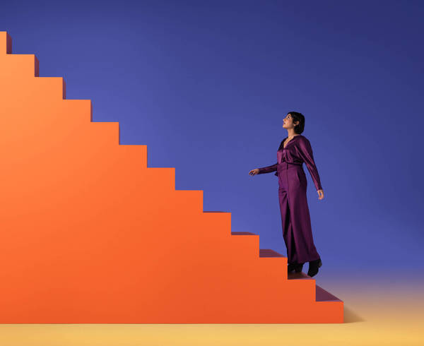 Woman in purple suit ascends bright orange staircase against vibrant blue backdrop, creating a striking, modern, and fashionable image.