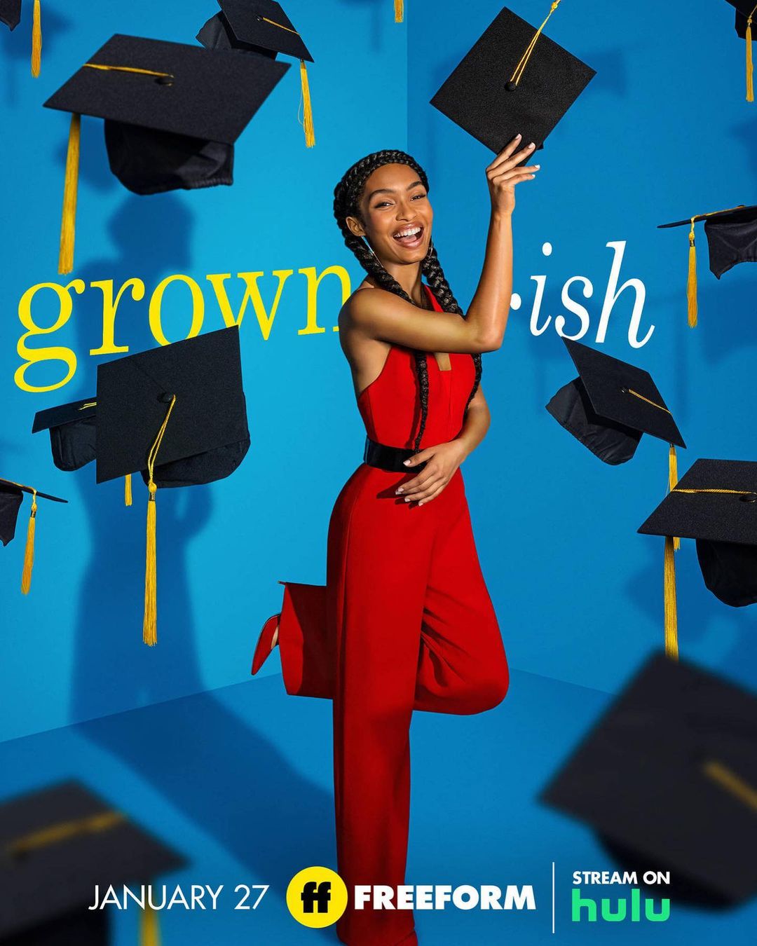Yara Shahidi in a red jumpsuit smiles in a promotional image for Grown-ish, surrounded by floating graduation caps on a blue backdrop.