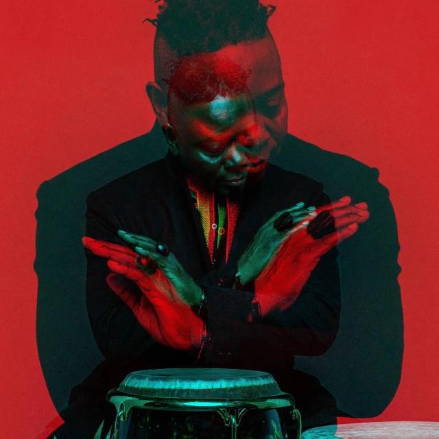 Portrait of a Black male musician seated behind a conga drum, with a green and red color shift and red background.