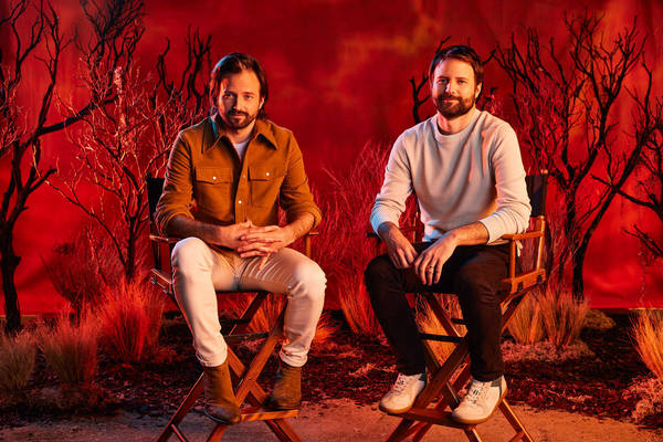 Two adult men sit on director's chairs in a photo studio with a red background and theatrical lighting for entertainment publicity.
