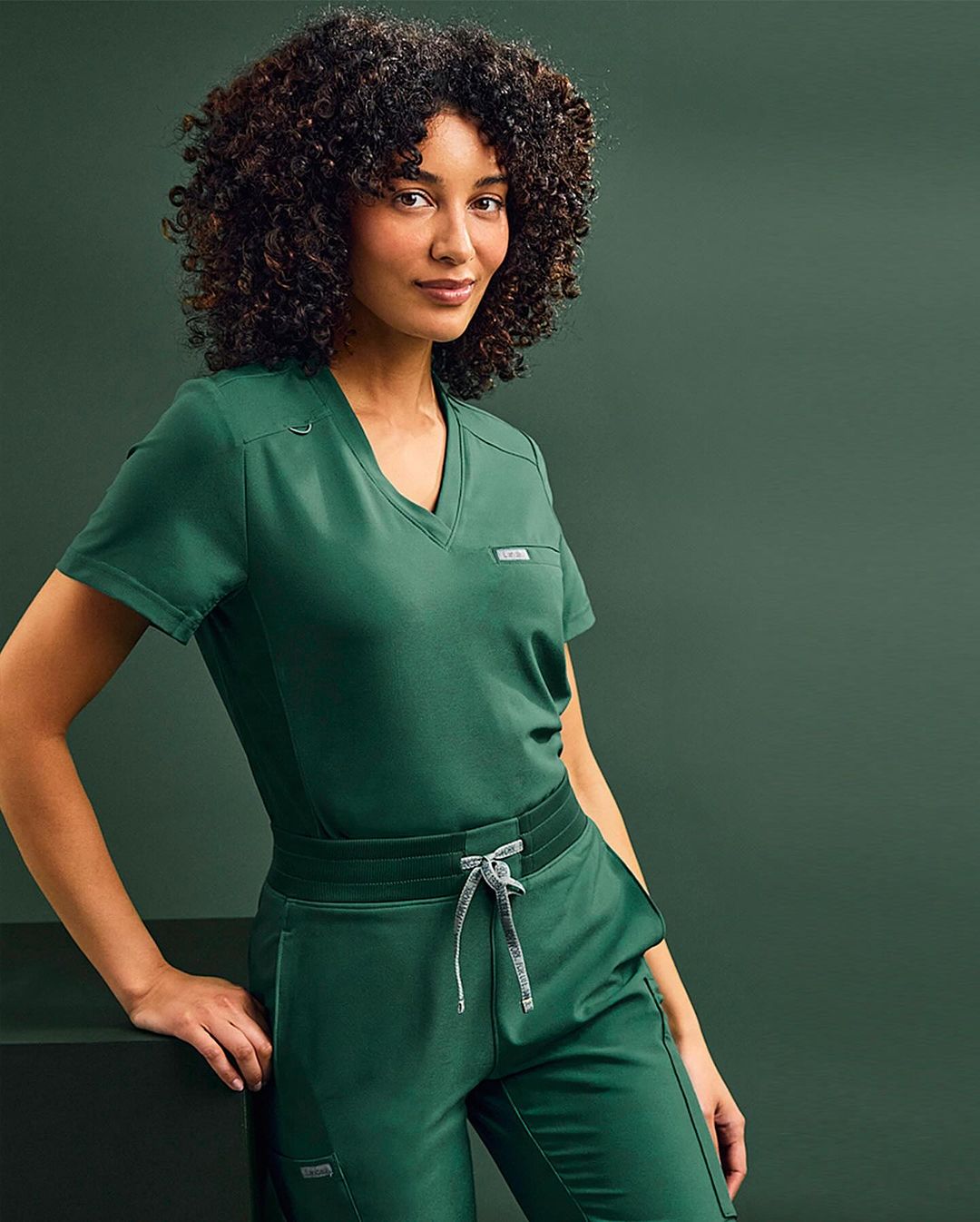Headshot of an adult woman with curly hair wearing green medical scrubs. She is leaning on a dark surface in a studio setting.