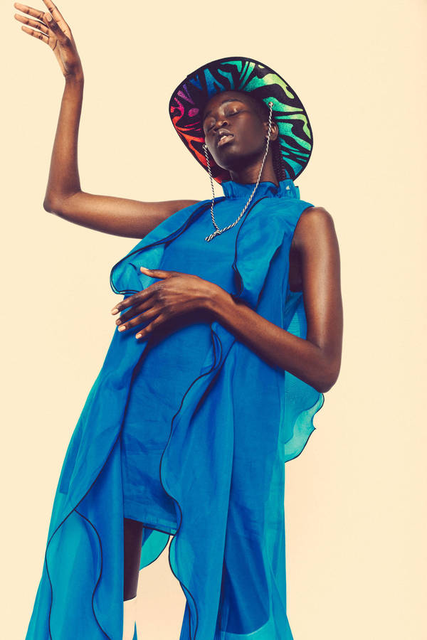 Elegant Black woman posing in a bright blue dress and colorful hat against a cream background, fashion editorial photography.