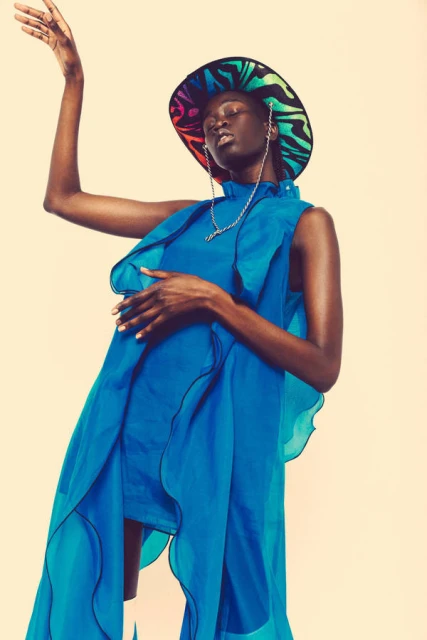 Elegant Black woman posing in a bright blue dress and colorful hat against a cream background, fashion editorial photography.