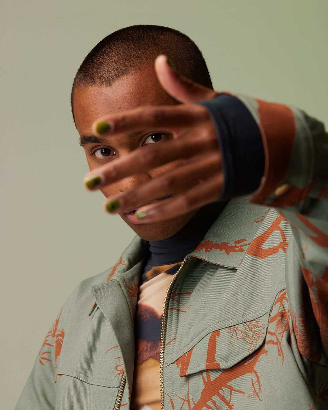 Close-up portrait of a young adult with green nail polish, wearing a patterned jacket, with hand partially covering their face.