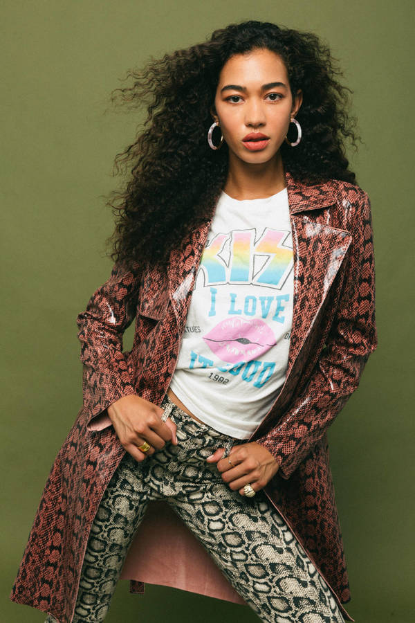 A young adult woman poses indoors wearing a Kiss band graphic t-shirt, snakeskin print jacket, and snakeskin print pants against a green backdrop.