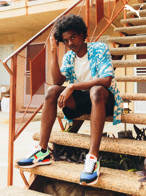 An adult male model sits on outdoor concrete stairs in a fashion editorial, wearing a tie-dye shirt, shorts, and multicolored Puma sneakers.