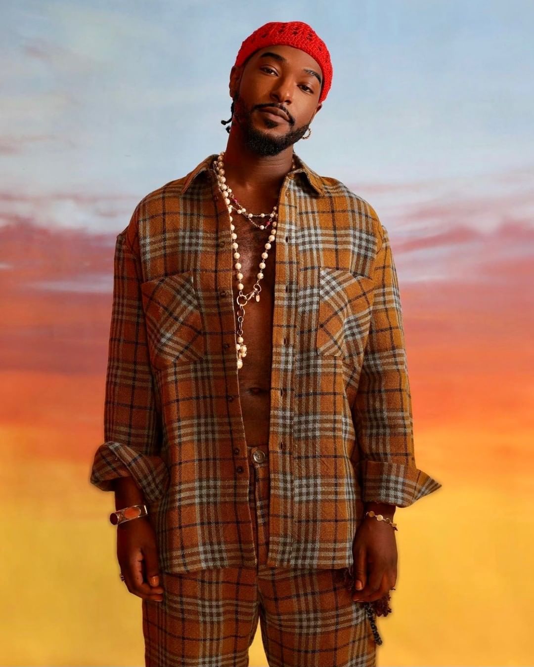 Adult male in a brown plaid outfit, wearing a red knit hat and pearl necklace, against a gradient sky background in a studio shot.
