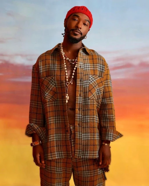 Adult male in a brown plaid outfit, wearing a red knit hat and pearl necklace, against a gradient sky background in a studio shot.