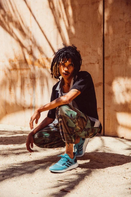 Young adult male with dreadlocks crouching outdoors in a black shirt, camo pants, and blue sneakers with a textured background.