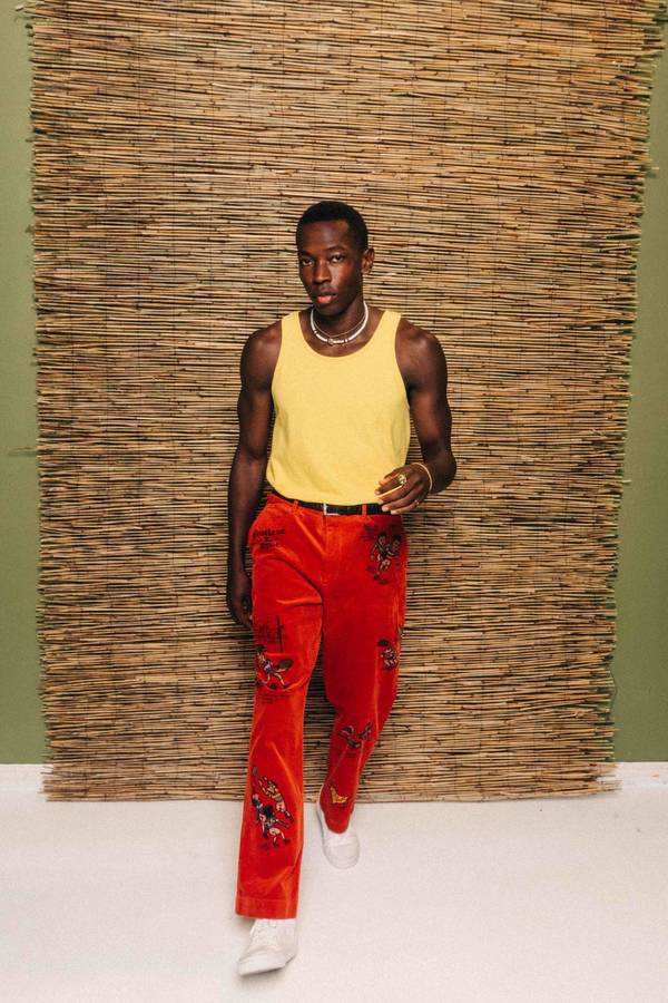 A fashionable Black male model stands in front of a woven bamboo background wearing a yellow tank top and floral embroidered orange pants.