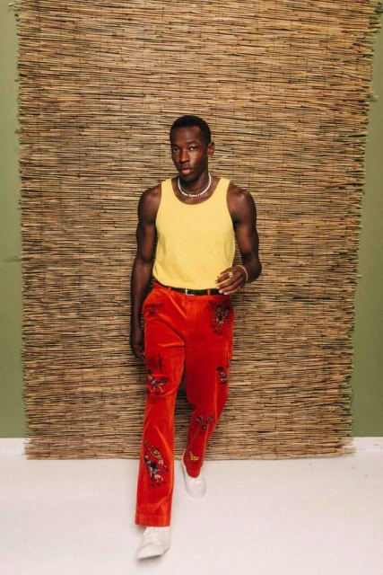 A fashionable Black male model stands in front of a woven bamboo background wearing a yellow tank top and floral embroidered orange pants.