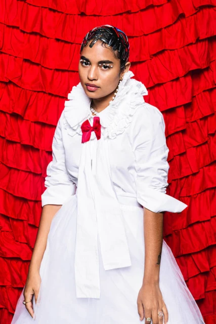 Woman in white shirt dress with red bow against red frilled backdrop. Close-up fashion portrait with pearl earrings and bold eye makeup.