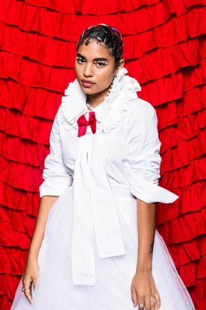 Woman in white shirt dress with red bow against red frilled backdrop. Close-up fashion portrait with pearl earrings and bold eye makeup.