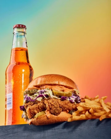 Close-up of a fried chicken sandwich with coleslaw and pickles, accompanied by a bottle of orange soda and a side of french fries.
