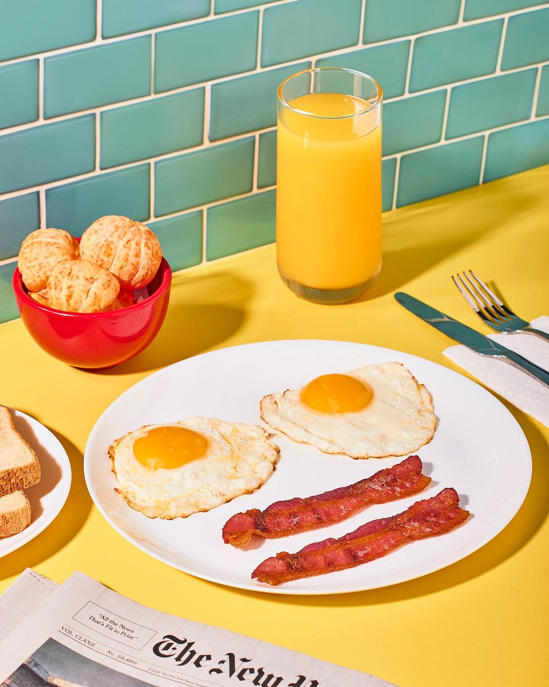 Overhead shot of breakfast with fried eggs, bacon, orange juice, a bowl of tangerines, and a newspaper on a yellow table.