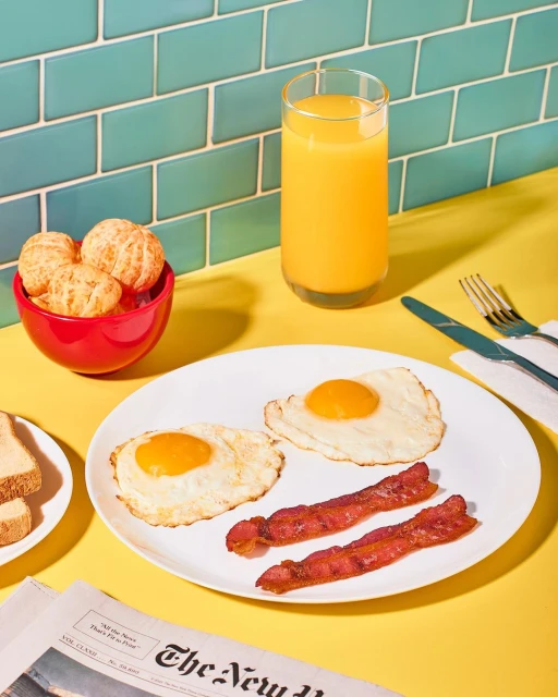 Overhead shot of breakfast with fried eggs, bacon, orange juice, a bowl of tangerines, and a newspaper on a yellow table.