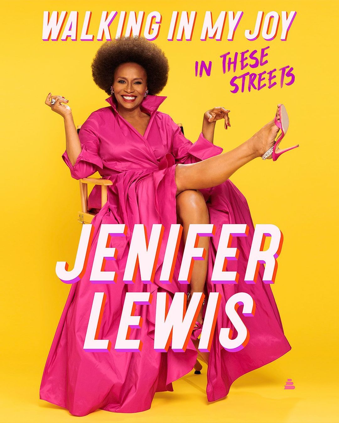 Jenifer Lewis sits in a director's chair with her leg raised, wearing a magenta dress and pink high heels, against a yellow backdrop.