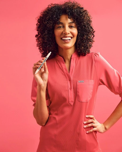 Smiling woman in coral top holds pen against pink backdrop. Studio portrait highlights curly hair, bright expression, and healthcare apparel.
