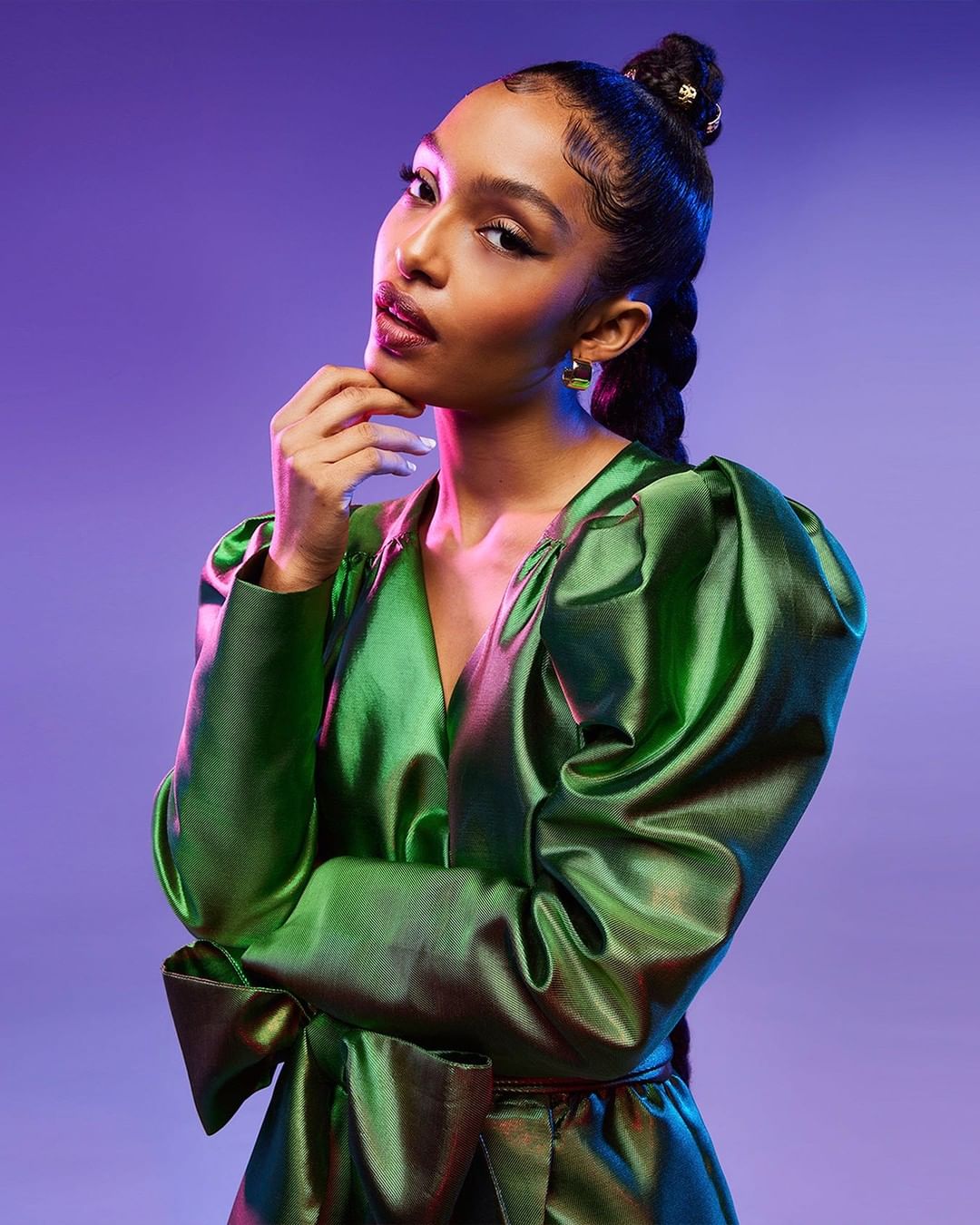 A fashionable young Black woman poses in a shiny green dress with puffed sleeves against a purple background, in a beauty style portrait.