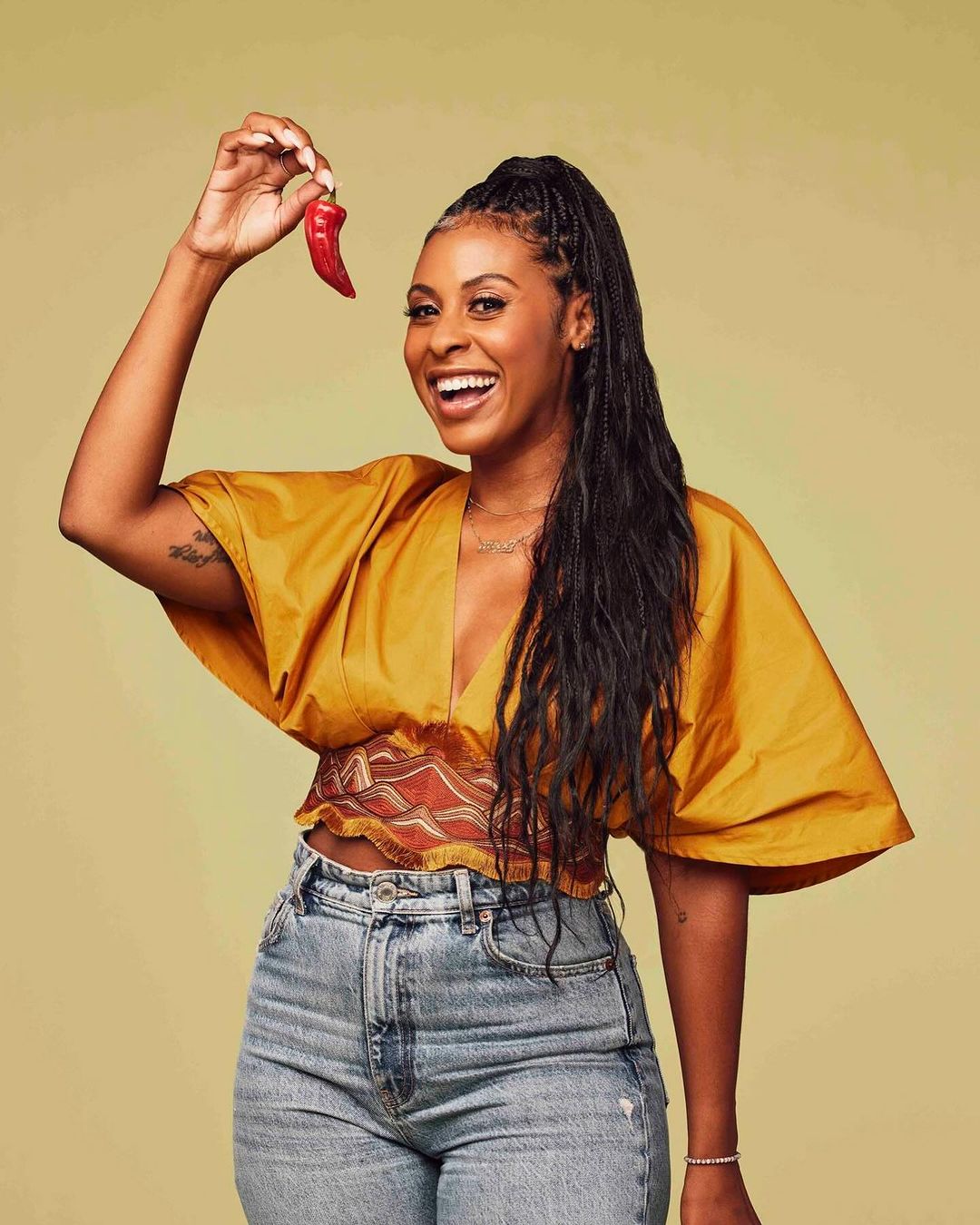 A smiling Black woman in a mustard top holds a red chili pepper, highlighted against a pale green background in a bright, eye-level studio shot.