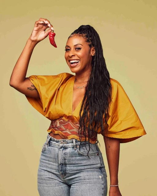A smiling Black woman in a mustard top holds a red chili pepper, highlighted against a pale green background in a bright, eye-level studio shot.