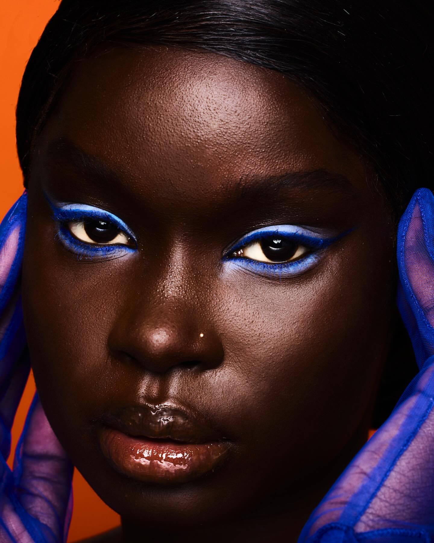 Close-up portrait of a dark-skinned woman with blue eye makeup and sheer blue gloves against an orange backdrop. Beauty and fashion.