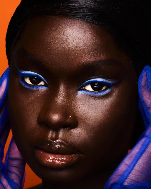 Close-up portrait of a dark-skinned woman with blue eye makeup and sheer blue gloves against an orange backdrop. Beauty and fashion.