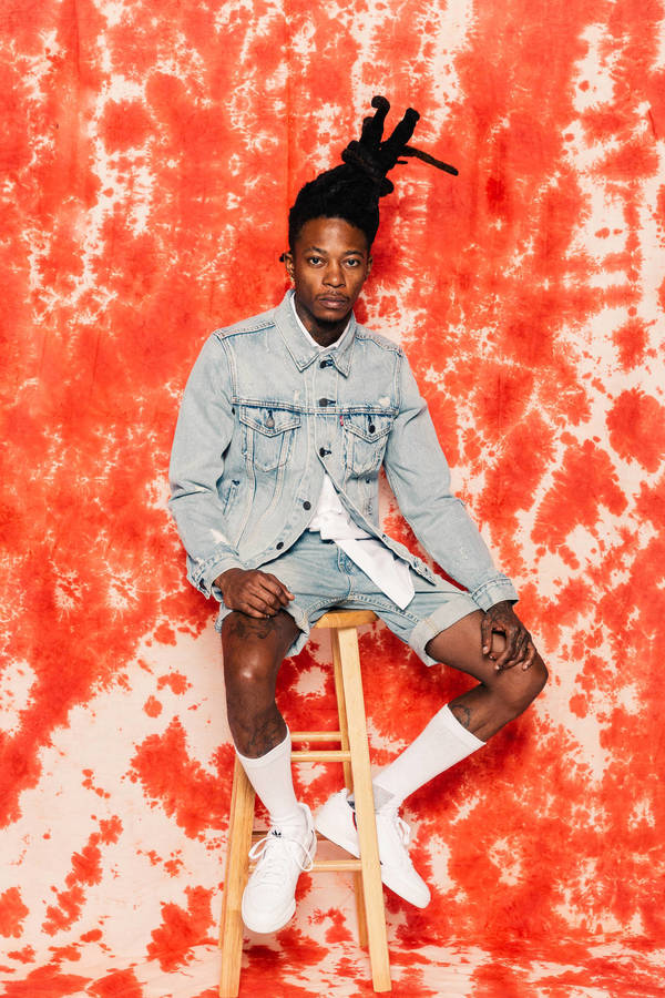 A young adult with dreadlocks is sitting on a wooden stool against a red and white tie-dye backdrop, wearing a denim jacket and white shoes.