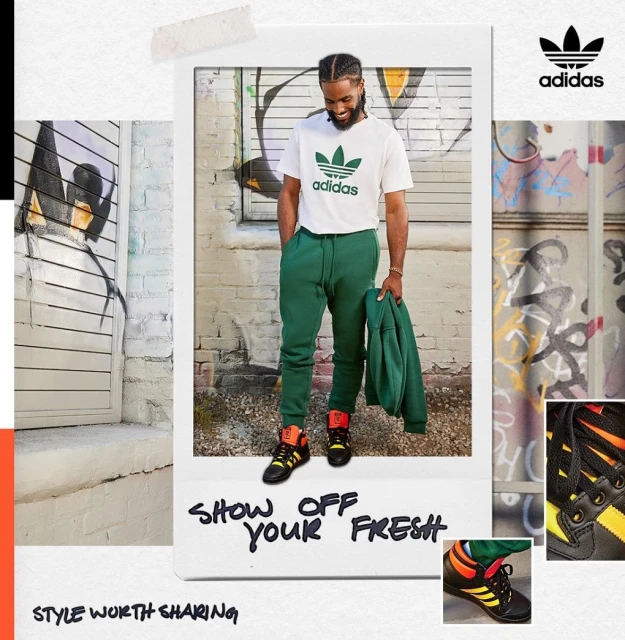Man in a white Adidas logo t-shirt and green sweatpants posing in front of a graffiti wall, styled with black and orange sneakers. 