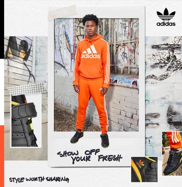 Young man in orange Adidas tracksuit poses indoors against a stylized wall. He wears black sneakers and the Adidas logo is visible.