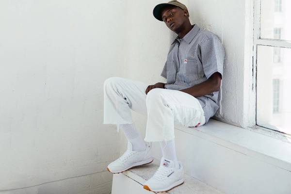 Young adult Black male wearing striped button-up shirt, white pants and sneakers sitting inside next to a window with natural light.