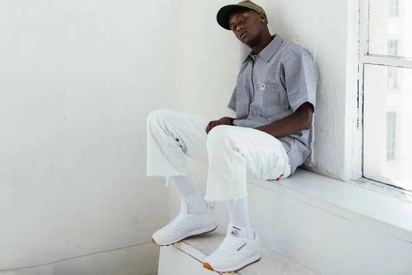 Young adult Black male wearing striped button-up shirt, white pants and sneakers sitting inside next to a window with natural light.
