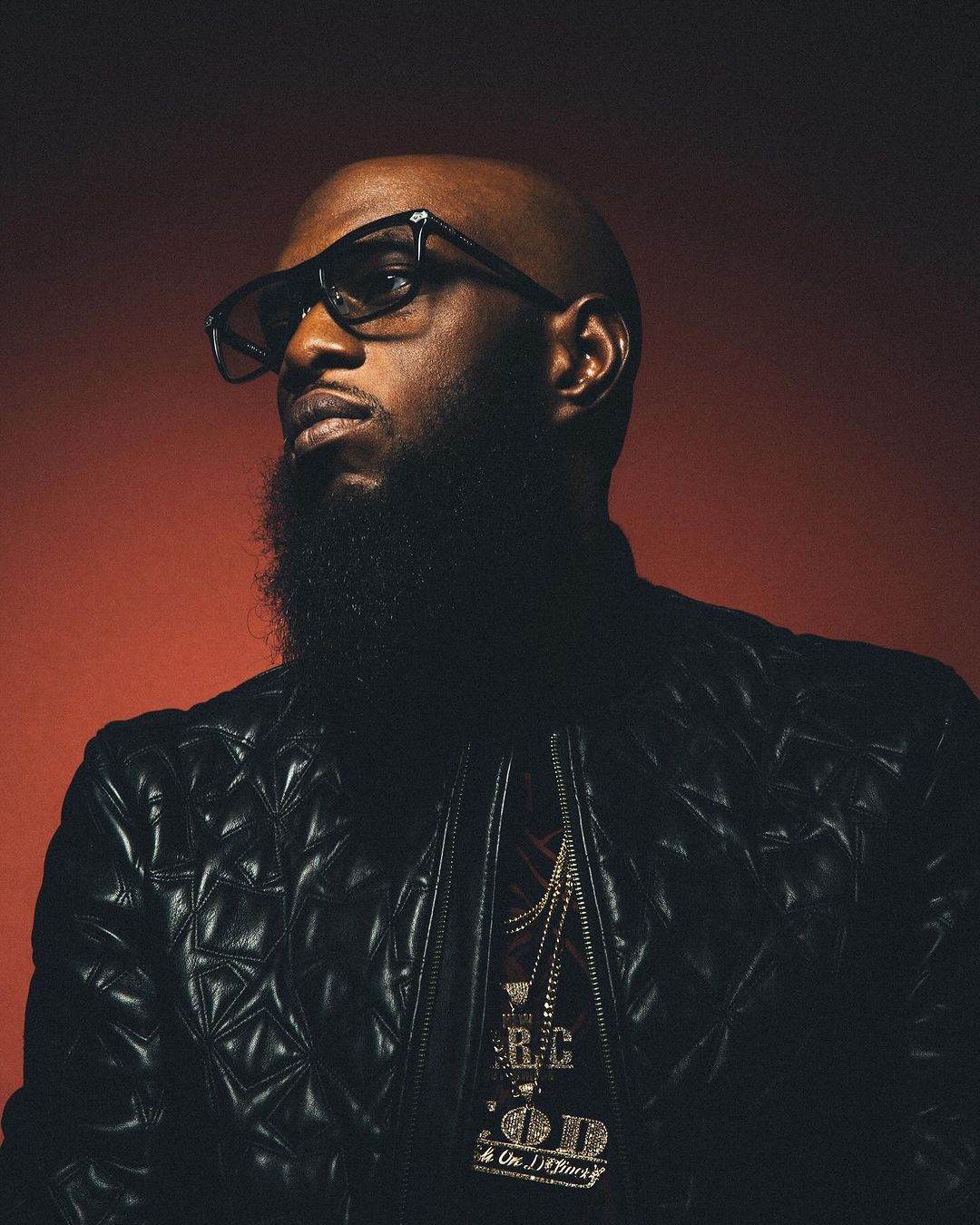Close-up portrait of a bald adult Black man with a beard wearing glasses, a leather jacket, and gold chain necklaces against red backdrop.