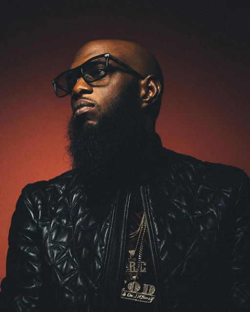 Close-up portrait of a bald adult Black man with a beard wearing glasses, a leather jacket, and gold chain necklaces against red backdrop.