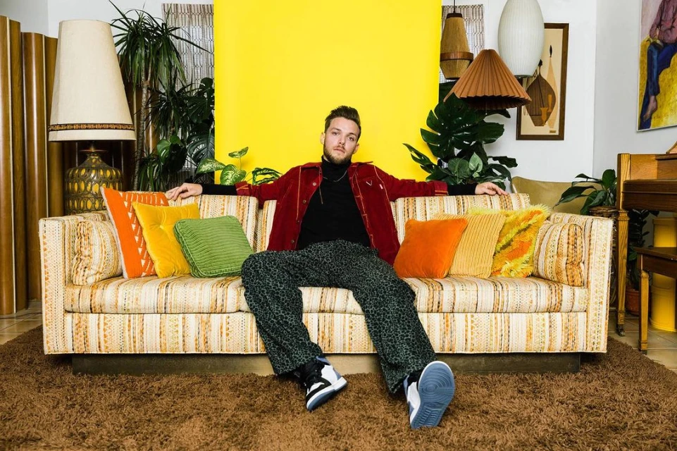 Young man with beard and patterned pants poses on a retro striped sofa with throw pillows indoors in front of a bright yellow wall.