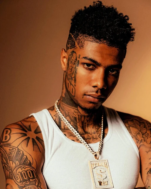Portrait of rapper Blueface wearing a white tank top and diamond jewelry, showcasing tattoos on his face, neck, and arms against a brown background.