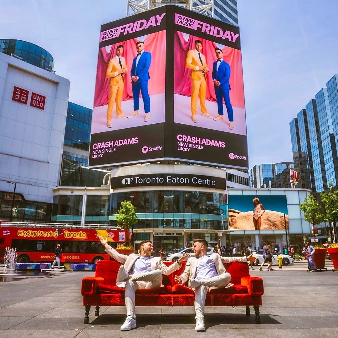 Two adult men sit on a red velvet couch in a city square, promoting music billboard for Crash Adams in Toronto, Canada on a sunny day.