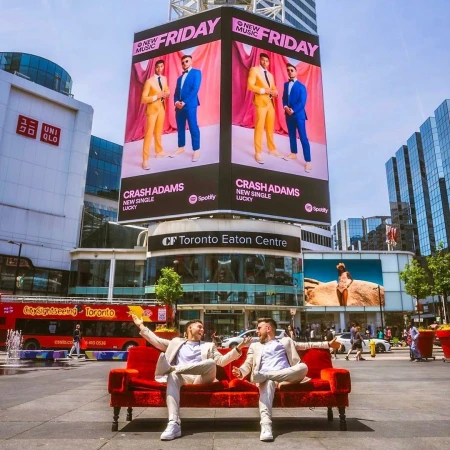 Two adult men sit on a red velvet couch in a city square, promoting music billboard for Crash Adams in Toronto, Canada on a sunny day.