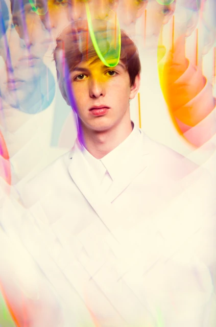 Portrait of a young adult male in a white suit jacket against a bright background with blurred, colorful, abstract shapes.