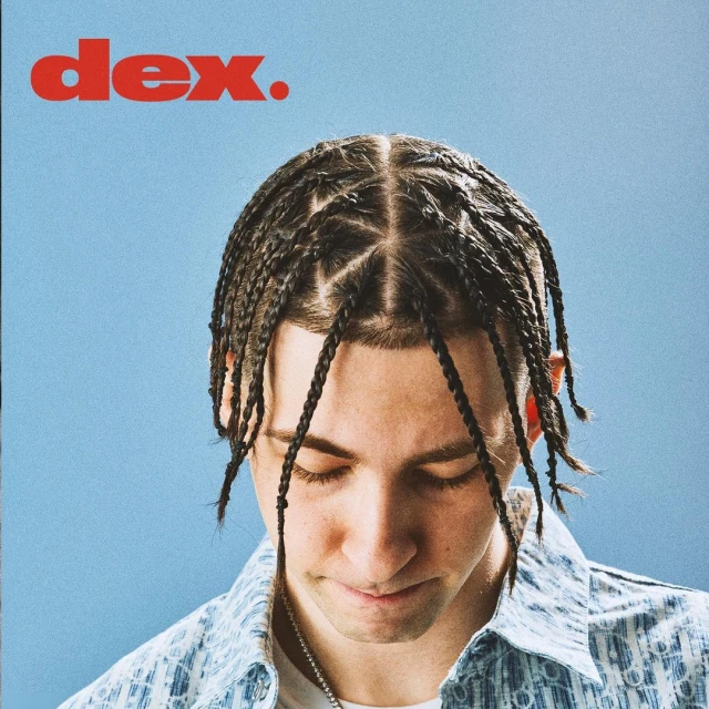 Close-up portrait of a young adult male with braided hair against a light blue background and red text in the upper left corner.