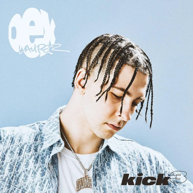 Portrait of rapper and songwriter, Gume, posing in front of a light blue background while wearing a printed jacket and silver chain.