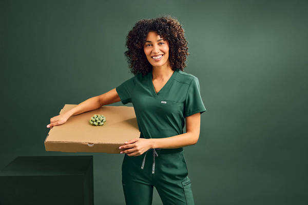 Smiling woman in green medical scrubs holds a pizza box with a succulent on top in a studio with a dark green background.