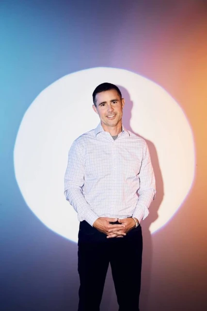 Portrait of a smiling adult man in a button-down shirt, standing in front of a white circle against a gradient blue and orange background.