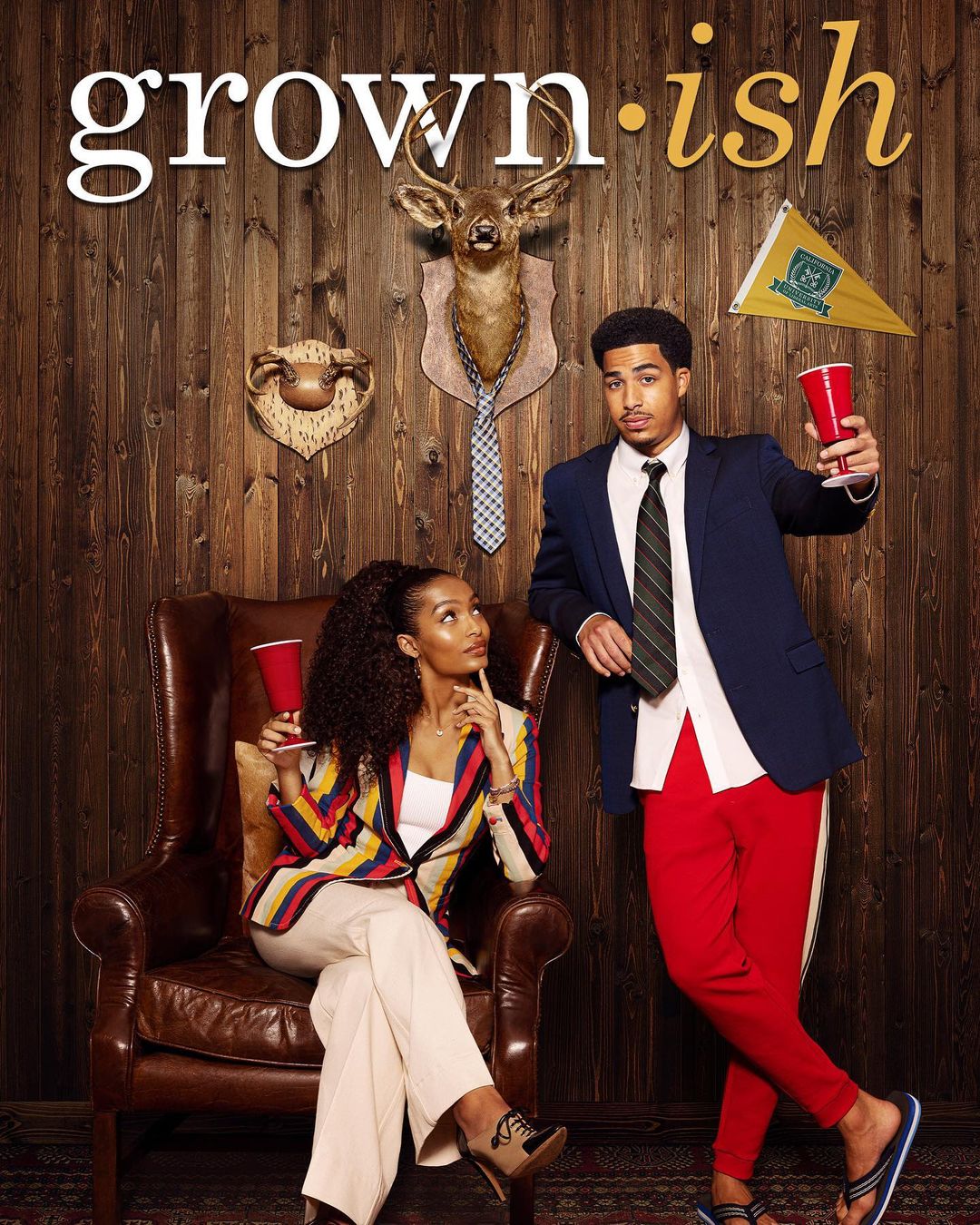 Promotional shot of Yara Shahidi in an armchair and Trevor Jackson standing, against a wood-paneled wall for 'Grown-ish'.