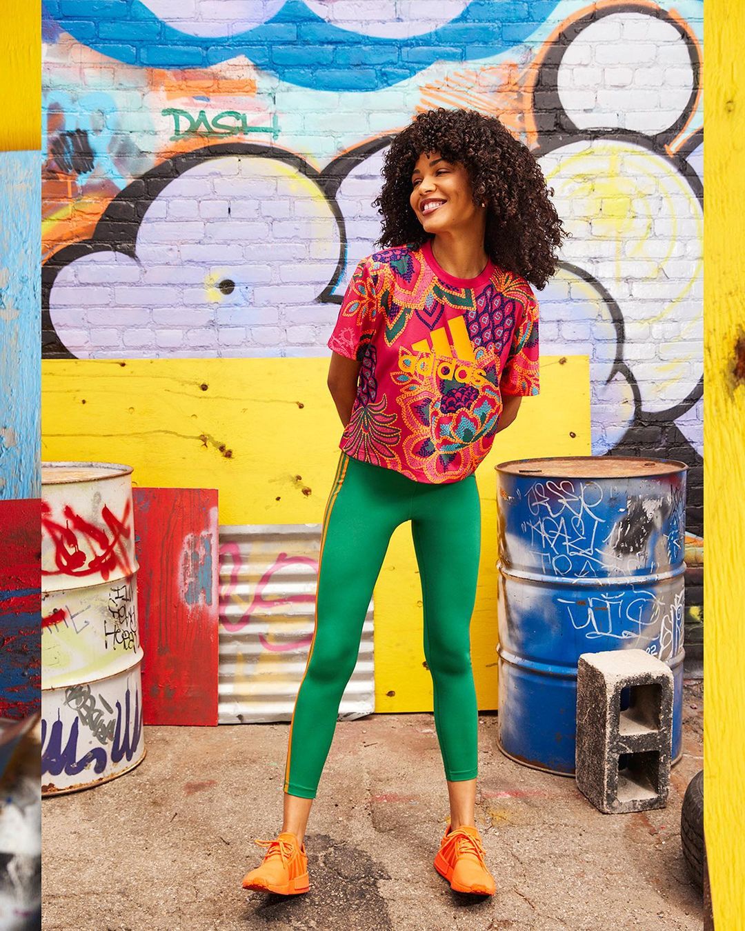 Smiling African-American woman in sportswear poses in front of urban graffiti art, wearing a colorful top, green leggings, and orange sneakers.